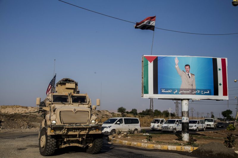 FILE - In this Saturday, Oct. 26. 2019 file photo, a U.S. military vehicle drives south of the northeastern city of Qamishli, likely heading to the oil-rich Deir el-Zour area where there are oil fields, or possibly to another base nearby, as it passes by a poster showing Syrain President Bashar Aassad. President Donald Trump's decision to dispatch new U.S. forces to eastern Syria to secure oil fields is being criticized by some experts as ill-defined and ambiguous. But the residents of the area, one of the country's most remote and richest regions, hope the U.S. focus on eastern Syria would bring an economic boon and eliminate what remains of the Islamic State group. (AP Photo/Baderkhan Ahmad, File)